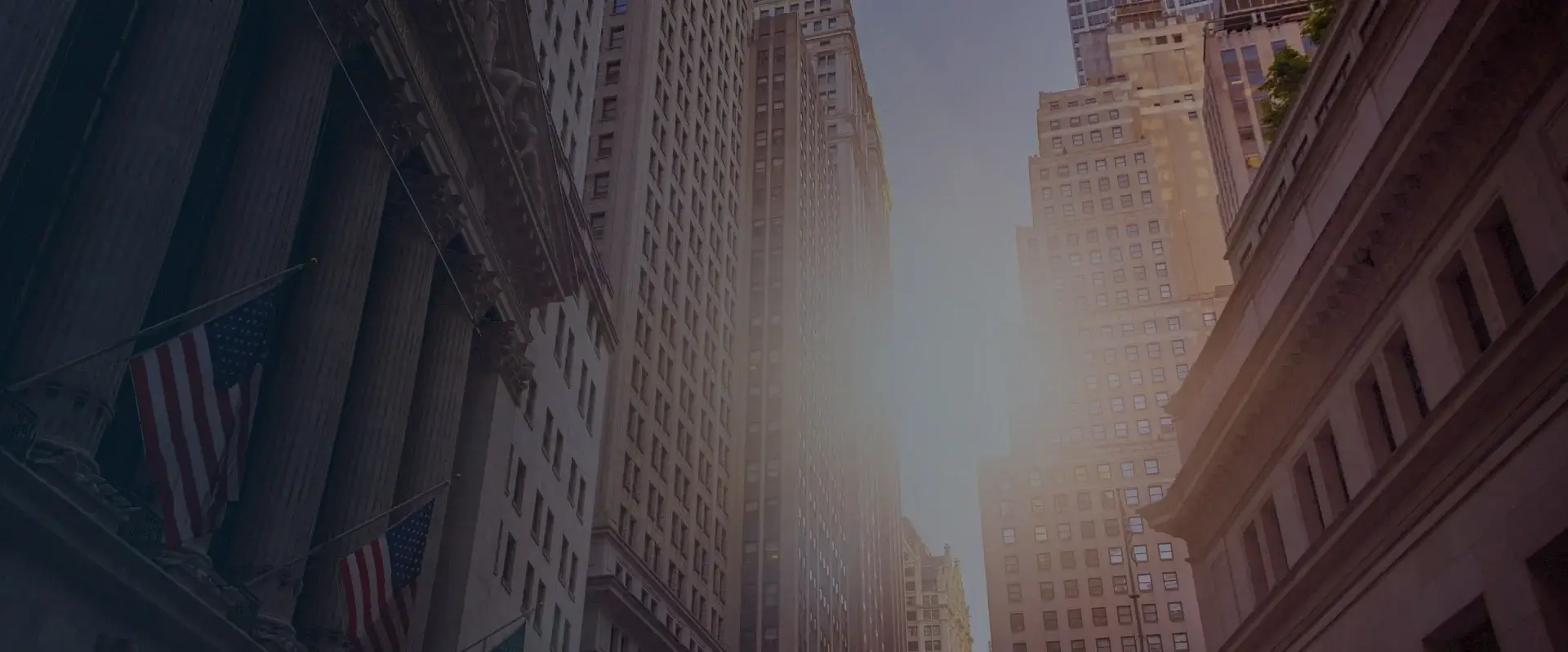 Sunlit street view of tall skyscrapers and a building with American flags on the left side.