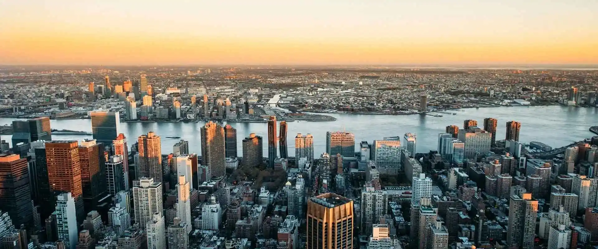 Aerial view of a city skyline with tall buildings by a river at sunset, with a golden sky in the background.