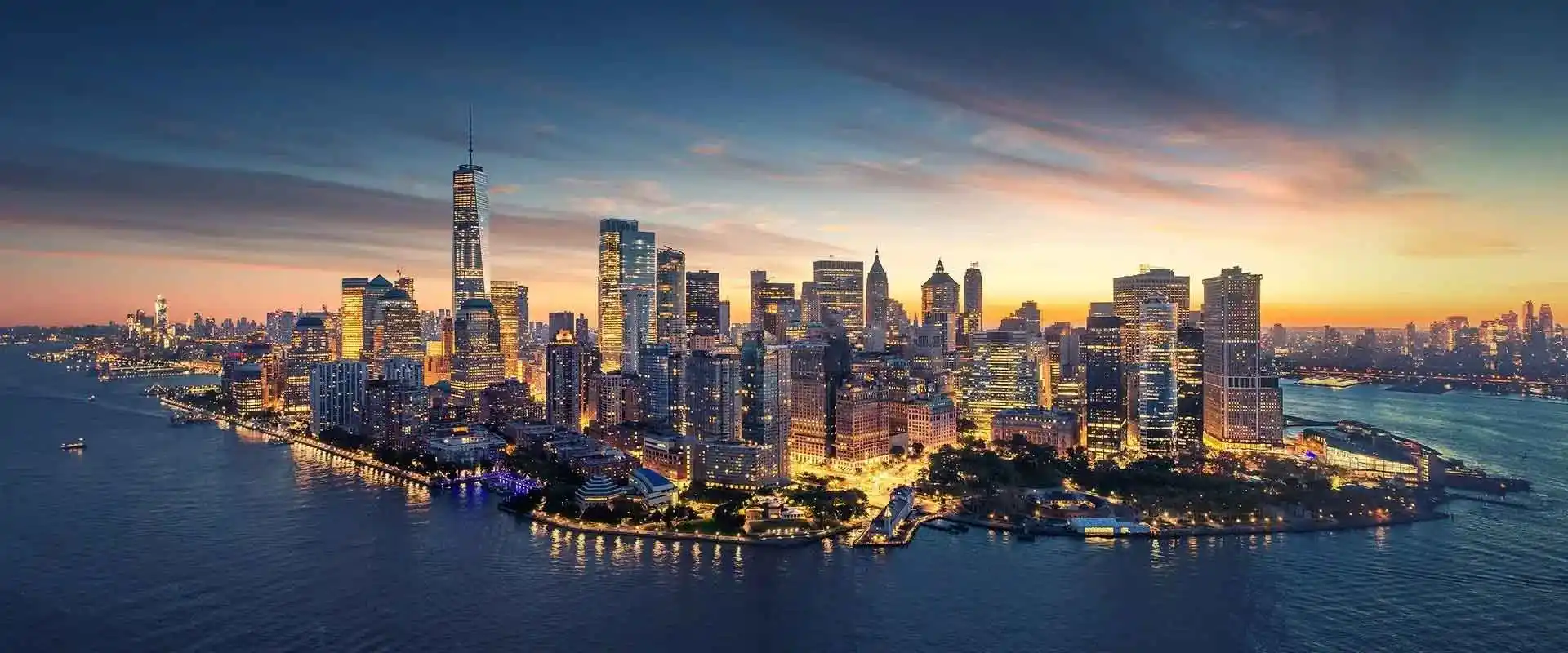 Aerial view of skyline at dusk with illuminated skyscrapers and waterfront along the Hudson River.