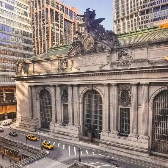 Grand Central Terminal facade with large clock and statues, surrounded by tall city buildings and yellow taxis on the street below.