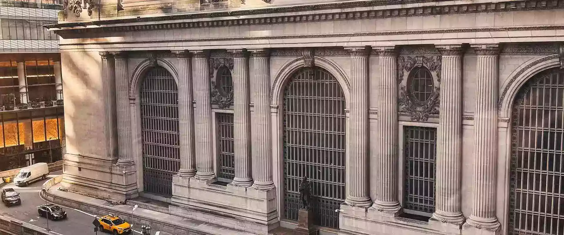 Exterior view of a grand neoclassical building with tall columns, large arched windows, and cars passing by on the street below.
