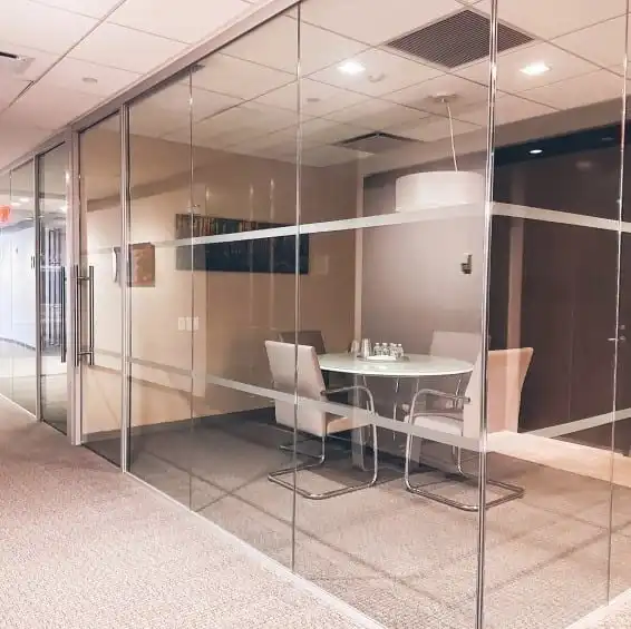 Modern conference room with glass walls, featuring a round table, four chairs, and a hanging light. The room conveys a clean and professional tone.